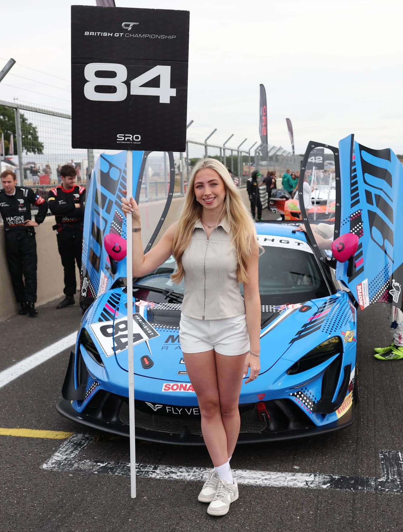 A grid girl holding an SRO board beside the number 84 Mahiki Racing McLaren Artura GT4 on the grid