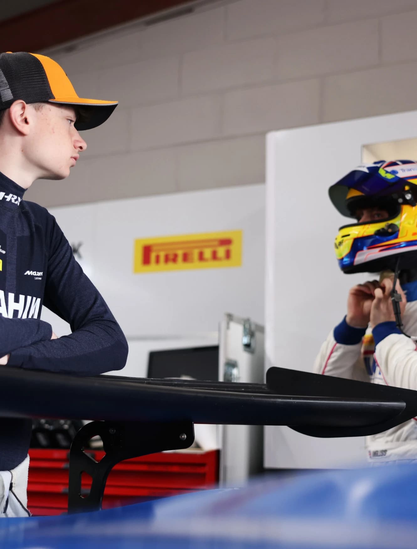 Revie Lake leaning on the car's rear wing in the garage as another driver pulls on a helmet