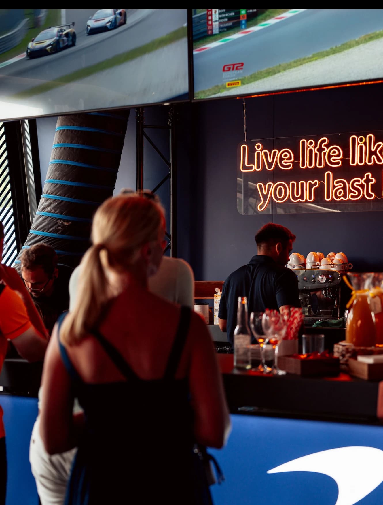 Guests at the hospitality bar under a neon sign reading Live life like it's your last lap