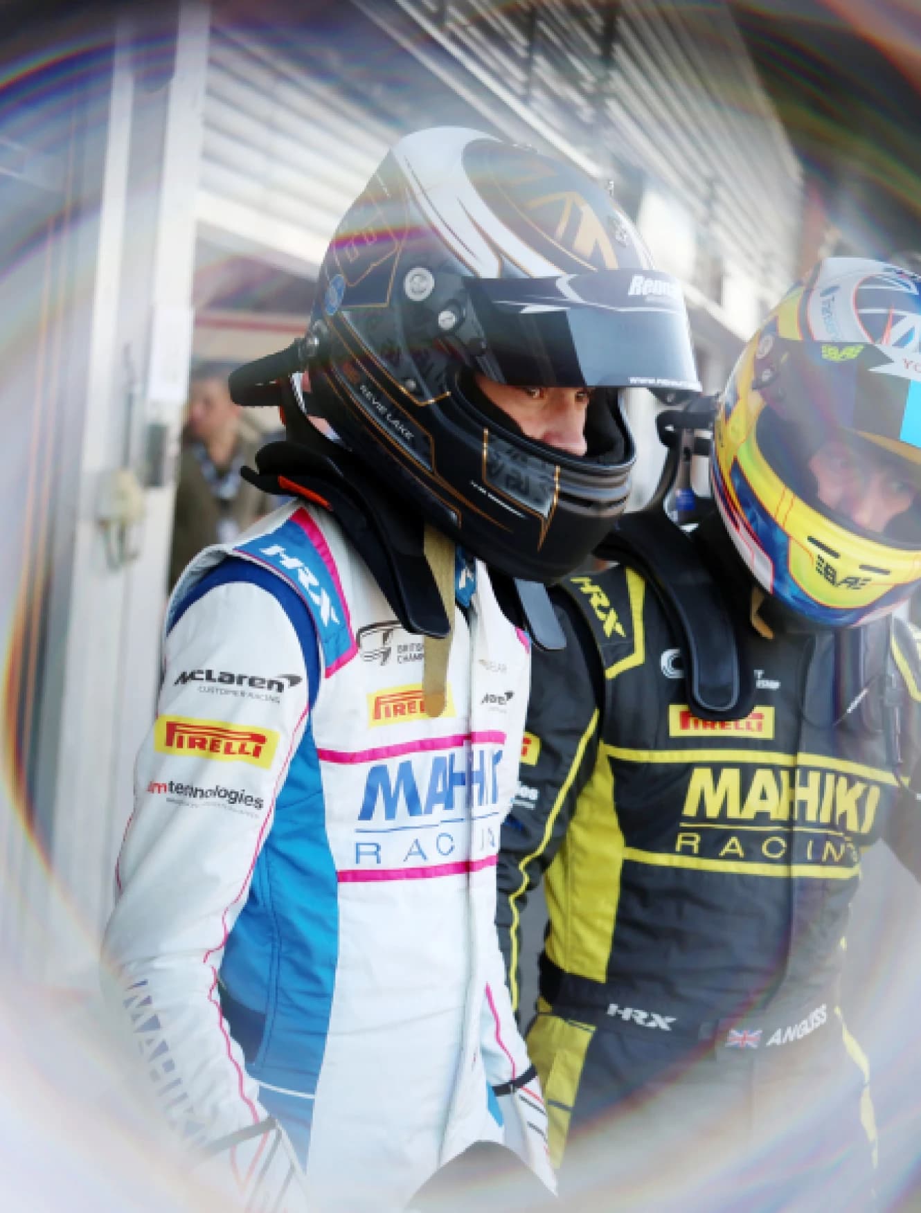 Two Mahiki Racing drivers in helmets and race suits waiting in the pit lane