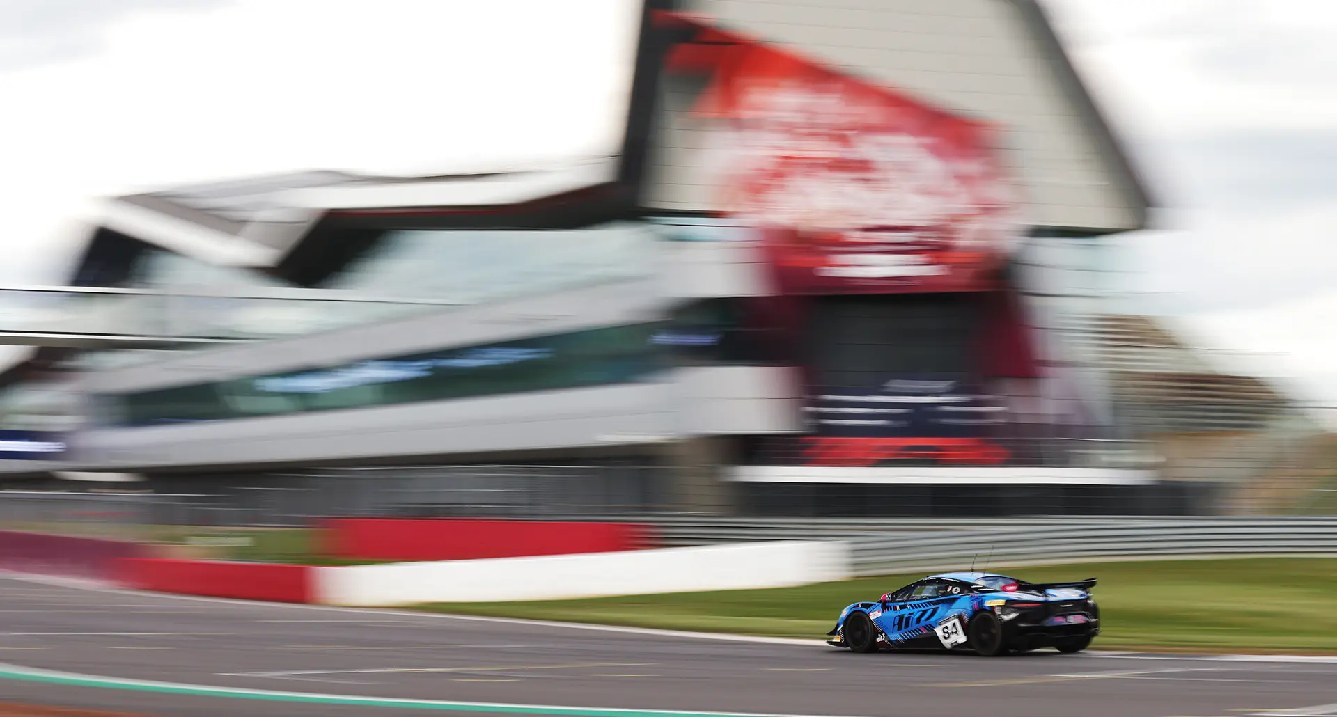 The number 84 Mahiki Racing McLaren Artura GT4 panning past the Silverstone Wing pit building